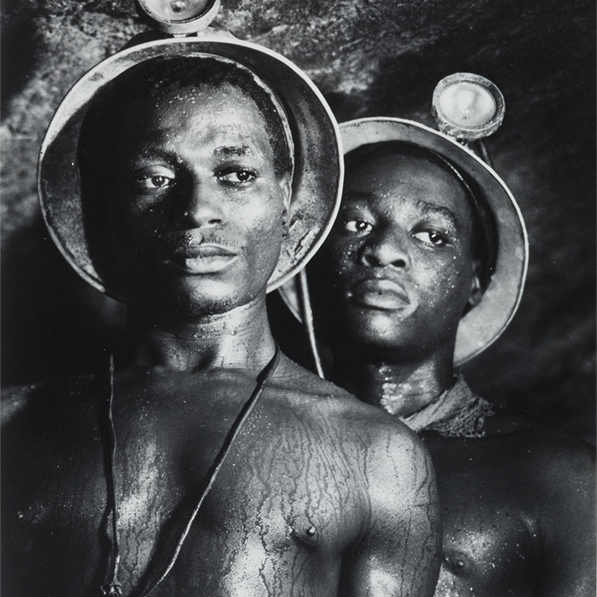 Black and white photo of two black men in a mine. They are shirtless and wearing miner's helmets.